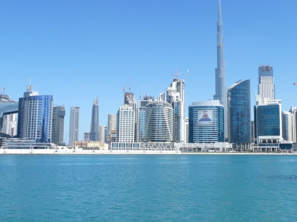 Aerial split-view comparison of Downtown Dubai Burj Khalifa and Dubai Fountain against Business Bay’s canal waterfront and modern high-rise towers, highlighting the urban contrast between luxury tourism and business living.
