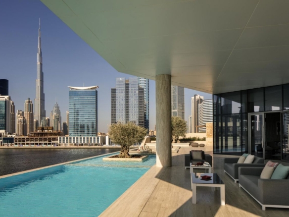 A smiling mixed-race couple holding golden house keys and a city blueprint, standing on a balcony overlooking Dubai's skyline with the Burj Khalifa in the background, ready to buy property in Dubai as foreign investors.