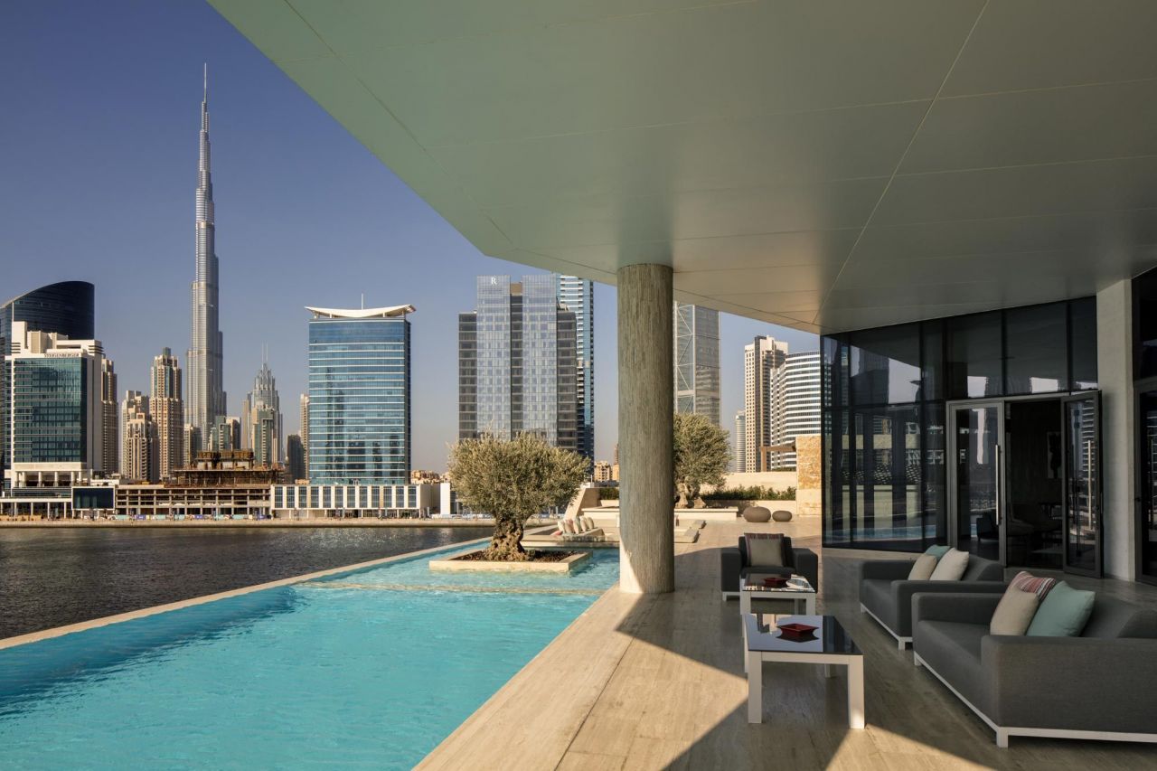 A smiling mixed-race couple holding golden house keys and a city blueprint, standing on a balcony overlooking Dubai's skyline with the Burj Khalifa in the background, ready to buy property in Dubai as foreign investors.
